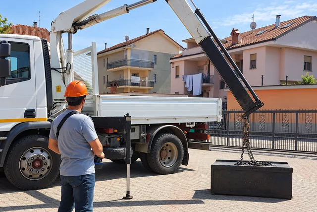 Immagine illustrativa rielaborata: operatore di spalle ai comandi di una gru su autocarro, con gancio e carico visibili.
