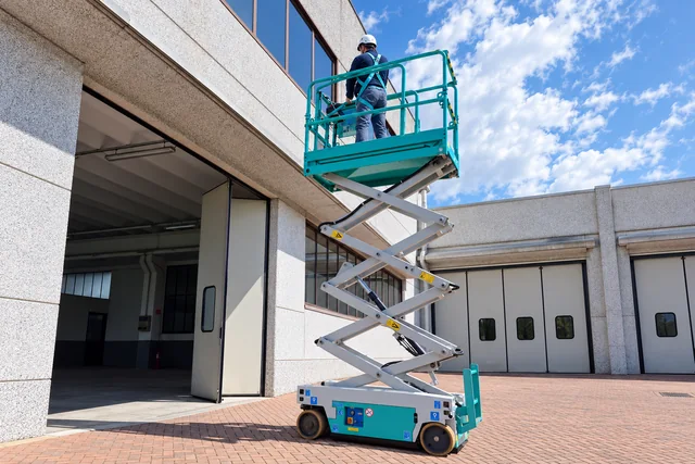 Immagine illustrativa rielaborata: operatore di spalle su una piattaforma elevabile a pantografo accanto a un edificio.