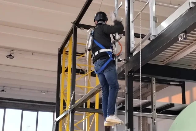 Foto reale ritoccata: operatore con casco e imbracatura durante una prova nell’area didattica per lavori in quota.